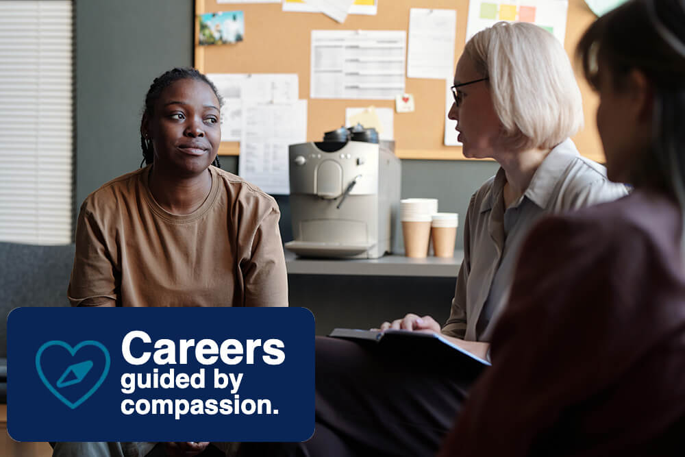 Three people are having a conversation in an office setting with a coffee machine and bulletin board in the background. A logo in the corner reads: Careers guided by compassion alongside a heart icon.