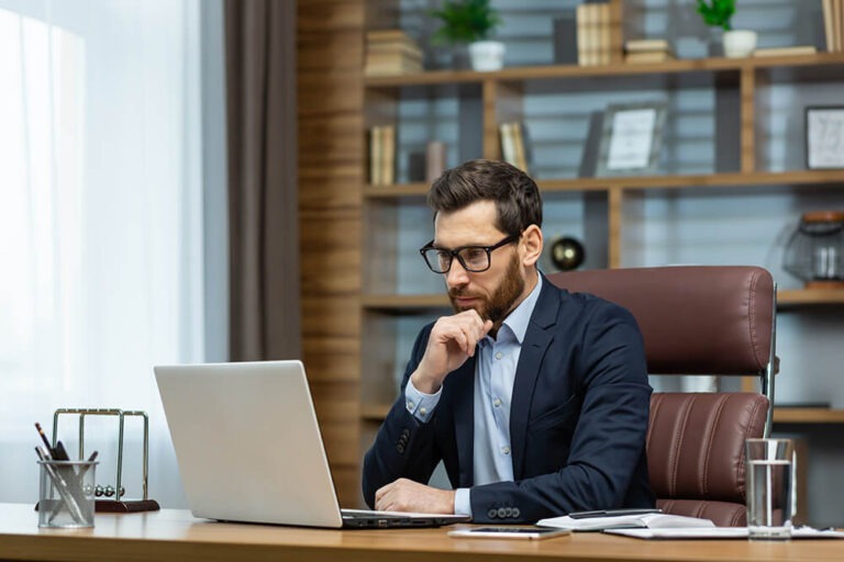 A man in a suit and glasses sits at a desk, concentrating on his laptop. The desk has a notebook, pen holder, and a glass of water. Books and decorative items fill the shelves in the background. Natural light comes through a window.